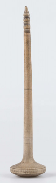 E/8019/ML - Przęślica krężołkowa z drewna brzozowego składa się z cienkiej deski – przysiadki, w której przy jednym końcu osadzona jest pionowa beleczka zw. piórem, z nasadzonym od góry ruchomym krężlem w kształcie wydłużonego stożka. Deseczka przysiadki o kształcie prostokątnym lekko zwężającym, od ok. połowy swej długości w kierunku pióra, i zakończonym głową zwierzęcą – psa. Pióro i krężel ozdobnie toczone. Krężel ma kształt stożka z rozszerzoną podstawą, w której znajduje się stożkowaty otwór, do osadzenia krężla na piórze. Dekoracja snycerska ryta znajduje się na zwężającej części przysiadki. Część zdobiona oddzielona małym stopniem. Grubość przysiadki w części zdobionej 2x większa od pozostałej części. Dekoracja snycerska tworzy ornament roślinny w postaci trzech ulistnionych drobno gałązek. Na zakończeniu ornamentu znajdują się ryte liście oraz inicjały „K.F.”. Głowa psa zdobiona drobnymi nacięcia. Zakończenie przysiadki wycięte faliście.