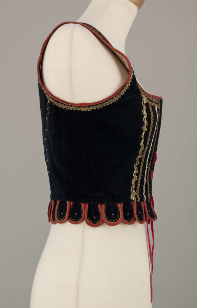 E/9558/ML - Gorset kobiecy z czarnego aksamitu, podszyty białą bawełnianą podszewką. Z przodu rozcięcie usztywnione fiszbinami, zaopatrzone w 10 drucianych haftek do przewlekania wstążeczki. Przód gorsetu ozdobiony złotym szychem, białymi tasiemkami i złotymi drobnymi cekinami naszytymi na nitki w ten sposób, że tworzą ornament w postaci falistej linii. Przód gorsetu zdobiony złotym i srebrnym szychem, klapki brzegi wykroju pachy wykończone czerwoną wełnianą tasiemką. Plecy w miejscach szwów ozdobione żółtą stebnówką i białymi koralikami. Od pasa gorsetu dookoła opadają klapki. Gorset szyty na maszynie.