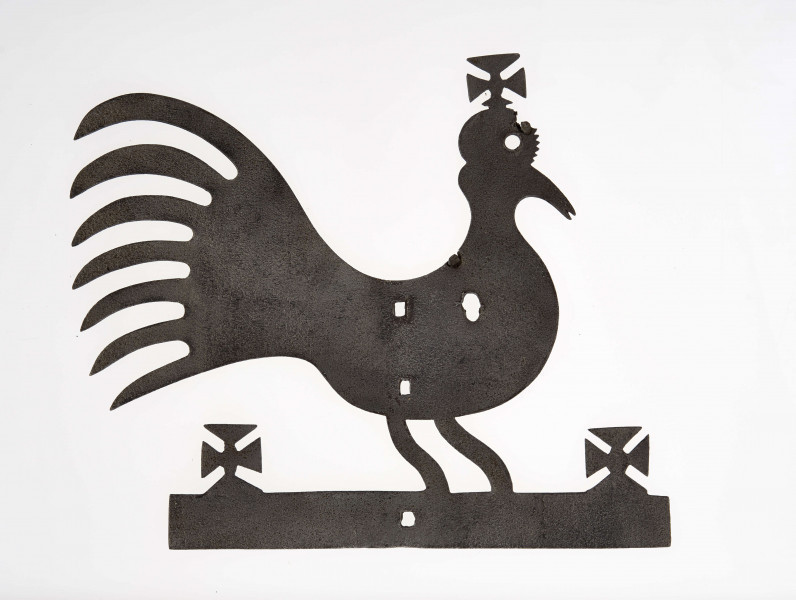 Kogut wykonany z żelaza techniką kucia. Stanowi zakończenie żelaznego krzyża zwieńczającego drewniany krzyż przydrożny. Posiada krzyżyki na głowie i w obu końcach podstawy.
