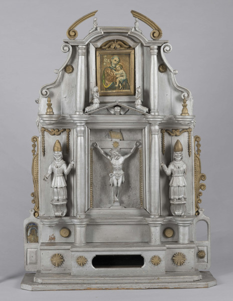 E/7373/ML - Ołtarzyk składa się z 3 części: podstawy i 2 pięter. Oba piętra podzielone na 3 pola (środkowe 24 cm, boczne 13,5 cm) przez kolumny. Podstawa posiadająca wysuniętą część środkową, ozdobiona jest 4 krzyżami greckimi wpisanymi w gwiazdę złot. Podstawę flankują 2 małe ołtarzyki: prawe brak przedstawienia, po lewej Madonna – złota postać na owalnym srebrnym polu, z granatową aureolą, nad nią na granatowym tle – 4 złote gwiazdki. Prawy ołtarzyk nad brakującym przedstawieniem ma złotą gwiazdkę. Lewemu jej brak. Górne pole podstawy zdobią 2 kolumienki oraz złote guzy pomiędzy. 
I piętro w części środkowej, pomiędzy kolumnami z tympanonem z głową aniołka i klęczącymi aniołami na daszku, postać Chrystusa Ukrzyżowanego w kolorze srebrnym, wraz ze złotymi  inskrypcją i pionowymi ząbkami. Po bokach, symetrycznie, przedstawienia  św. Mikołaja w złotej czapce biskupiej, na owalnej podstawie, z jedną ręką uniesioną , a druga opuszczoną trzymającą rękojeść. Ponad stylizowany złoty ornament roślinny. Całość piętra flankowana ornamentem przypominającym liście akantu i ślimacznice. 
II piętro w częściach bocznych zamknięte jest spływami wolutowymi , na polach po złotej kolumience/sterczynie i gwieździe. W części środkowej oleodruk ze św. Józefem z Dzieciątkiem, w złotej ramce. Całość wieńczą postaci ornatów w kolorze srebrnym i złote niepełne spływy wolutowe ze ślimacznicą. 
Brak wielu elementów. Plecy ołtarzyka stanowi 6 deseczek o szer. 10,5 cm i grubości 1,2 cm.