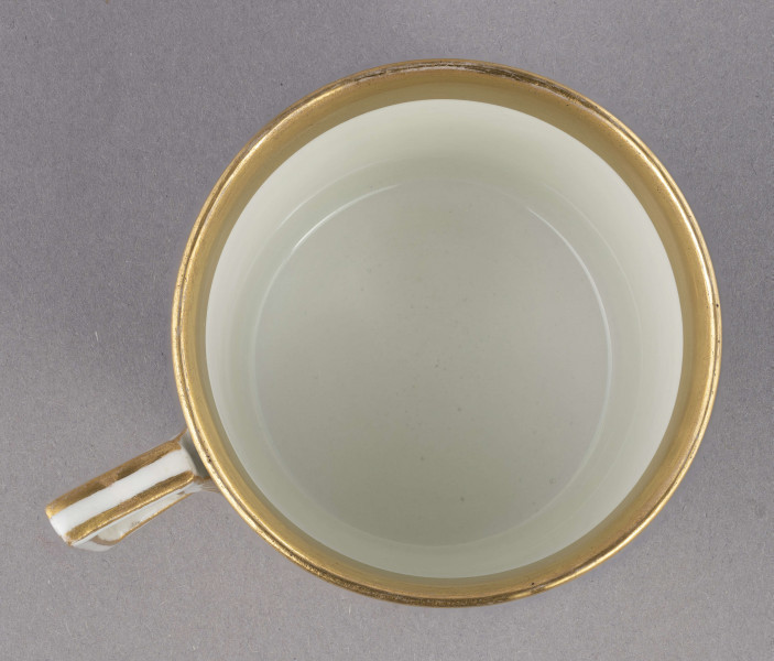 Filiżanka i spodek malowane w piórka. Filiżanka cylindryczna, ucho łamane z listkami w osadzeniach. Brzeg złocony obustronnie, złotem  konturowane ucho, złote kontury dzielą powierzchnię na ukośne pasy na przemian białe i jasnoszare. W nich malowane odpowiednio złote i czarne pióra.