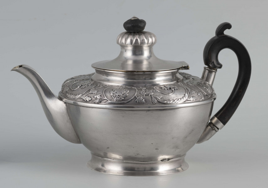 S/Mt/542/ML - Korpus w kształcie spłaszczonej kuli, na niskiej stopie, wylew w formie dzióbka, ucho drewniane malowane na czarno, osadzone w dwóch tulejkach, pokrywka na zawiasie, płaska, w części środkowej wypiętrzona i ozdobiona puklowaną rozetą.Górna część brzuśca ozdobiona sztancowaną taśmą z wzorem spiralnie zwinietej wici roślinnej z kwiatami, uchwyt przykrywki zakończony drewnianą rozetką.  
