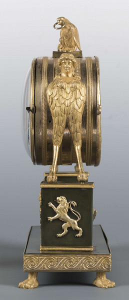 S/Mt/640/ML - Wsparty na czterech zwierzęcych łapach z brązu złoconego, podstawa prostokątna, zdobiona zgeometryzowanym fryzem, wyżej dwa prostokątne filary przechodzące w hybrydyczne figury podtrzymujące cylindryczną puszkę mechanizmu, zwieńczoną figurą orła. Tarcza emaliowana z podziałem arabskim - na niej sygnatura kursywą 