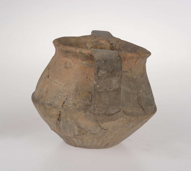 Naczynie wazowate, dwustożkowe z dwoma taśmowatymi uchami. Nad załomem brzuśca zdobienie w postaci linii rytych wypełnionych ukośnym kreskowaniem. Powierzchnia brunatna. Typ III/3 wg T. Liany.