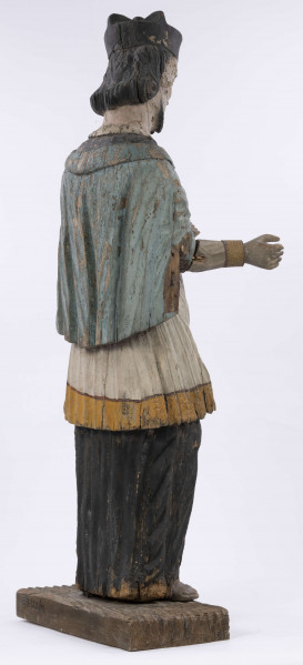 E/8422/ML - Rzeźba drewniana, pełna, polichromowana, przedstawiająca św. Jana Nepomucena. Zakomponowana frontalnie, z rękoma zgiętymi w łokciu i lekko rozłożonymi. Twarz owalna, pomalowana białą farbą. Małą broda i krótkie czarne wąsy. Nos krótki, prosty. Oczy owalne podkreślone czarnym konturem. Brwi łukowate, wąskie, czarne. Włosy czarne, ułożone w pukle. Na głowie czarny biret. Długa czarna sutanna, biała komża ozdobiona u dołu złotą koronką. Na piersiach krzyż, zawieszony na łańcuchu. Na komżę narzucona jasnoniebieska peleryna. Stopy lekko rozstawione. Obuty w sandały o jednym pasku. 

Konserwacja: 1990 r.