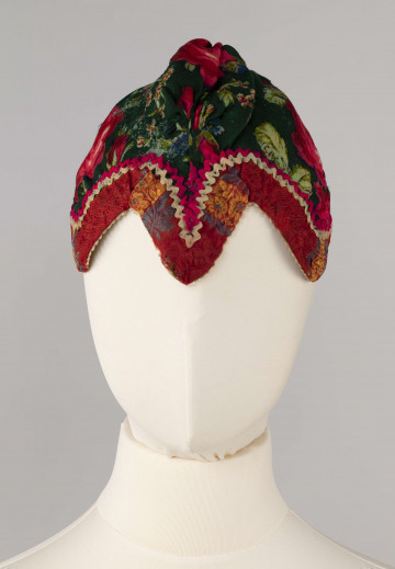 E/2121/ML - Czepek (kaptur) wykonany z wełnianej zielonej chusty szalinówki o motywach kwiatowych. Obszyty czerwoną wstążką, zdobioną motywami kwiatowymi w kolorze niebieskim i żółtym oraz białą ząbkowaną taśmą. Szew między wstążką a wzorzystym materiałem ozdobiony naszyciem taśmy pasmanteryjnej w kolorze białym i amarantowym. Nad czołem wycięty w ząb.