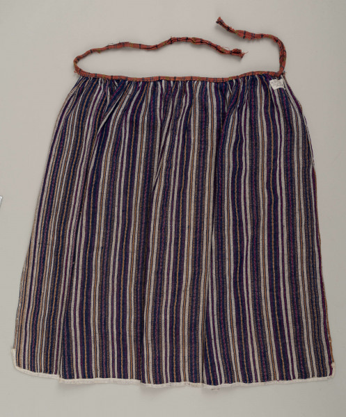 E/5997/ML - Fartuszek uszyty z jednego kawałka materiału - tkaniny samodziałowej bawełniano-wełnianej, tkanej splotem rypsowym w drobne paseczki w kolorze: białym, czerwonym, granatowym i bordo. Suto marszczony w oszewkę przechodzącą w troczki. Oszewka z materiału lnianego w kratkę czerwono-zielono-granatową. Dół obszyty tasiemką z białego bawełnianego płótna.