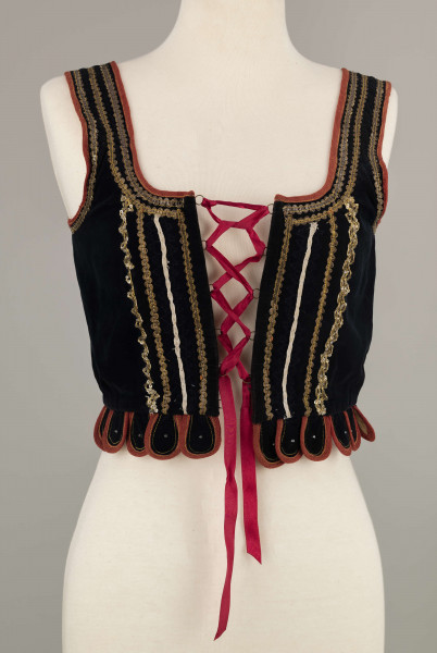 E/9558/ML - Gorset kobiecy z czarnego aksamitu, podszyty białą bawełnianą podszewką. Z przodu rozcięcie usztywnione fiszbinami, zaopatrzone w 10 drucianych haftek do przewlekania wstążeczki. Przód gorsetu ozdobiony złotym szychem, białymi tasiemkami i złotymi drobnymi cekinami naszytymi na nitki w ten sposób, że tworzą ornament w postaci falistej linii. Przód gorsetu zdobiony złotym i srebrnym szychem, klapki brzegi wykroju pachy wykończone czerwoną wełnianą tasiemką. Plecy w miejscach szwów ozdobione żółtą stebnówką i białymi koralikami. Od pasa gorsetu dookoła opadają klapki. Gorset szyty na maszynie.