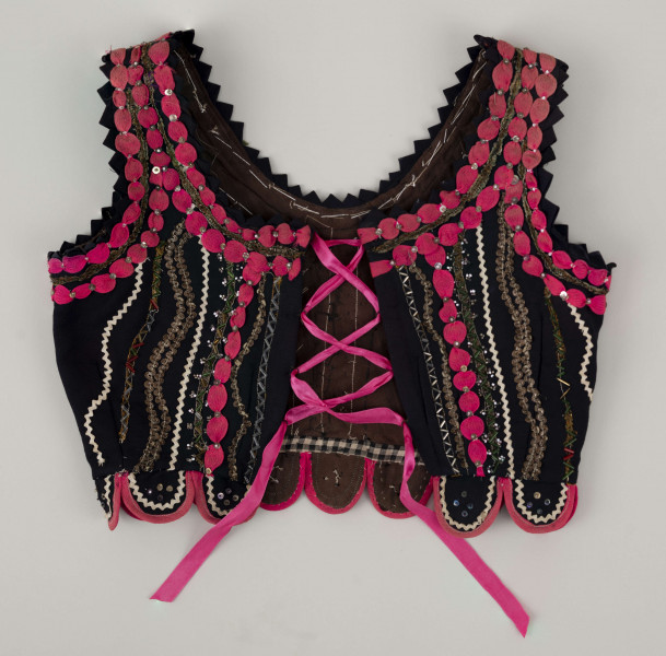 E/7358/ML - Gorset zszyty z 3 kawałków granatowej wełenki, podszyty brązową bawełnianą podszewką. Z przodu rozcięcie usztywnione fiszbinami, zaopatrzone w 10 haftek drucianych do przewlekania wstążeczki. Od pasa gorsetu dookoła opadają klapki. Cały gorset bogato zdobiony amarantowymi tasiemkami, żółtym i srebrnym szychem, białą krepiną, barwnymi koralikami i cekinami. Przód gorsetu, brzegi wykroju i pachy ozdobione dodatkowo granatowymi ząbkami. 