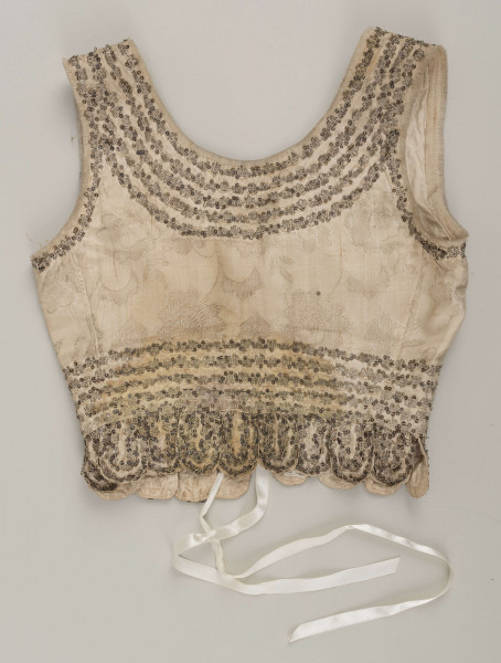 E/6235/ML - Gorset ślubny z białego wzorzystego atłasu. Składa się z 3 części: pleców i dwóch przodów. W pasie doszyte 17 owalnych klapek, zw. taszki. Wycięcie wokół szyi , przodów i rękawów wykończone białą wełnianą tasiemką, zw. szczotka. Całość zdobiona złotym szychem i srebrnymi cekinami. Podszyty białą bawełnianą podszewką.  