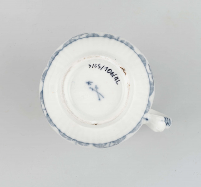 Dzbanek gruszkowaty na niskiej pierścieniowatej podstawie, ucho wolutowe, wylew trókątny, powierzchnia drobno kanelowana. Malowany podszkliwnie błękitem kobaltowym we wzór suchych kwiatów (kocanka), poniżej brzegu górnego na zewnatrz i wewnątrz malowana cienka obwódka, na dnie brzegu uszka gałązka. 
