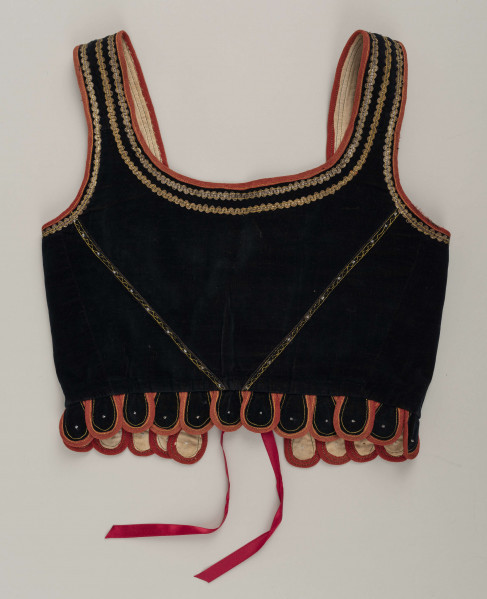 E/9558/ML - Gorset kobiecy z czarnego aksamitu, podszyty białą bawełnianą podszewką. Z przodu rozcięcie usztywnione fiszbinami, zaopatrzone w 10 drucianych haftek do przewlekania wstążeczki. Przód gorsetu ozdobiony złotym szychem, białymi tasiemkami i złotymi drobnymi cekinami naszytymi na nitki w ten sposób, że tworzą ornament w postaci falistej linii. Przód gorsetu zdobiony złotym i srebrnym szychem, klapki brzegi wykroju pachy wykończone czerwoną wełnianą tasiemką. Plecy w miejscach szwów ozdobione żółtą stebnówką i białymi koralikami. Od pasa gorsetu dookoła opadają klapki. Gorset szyty na maszynie.