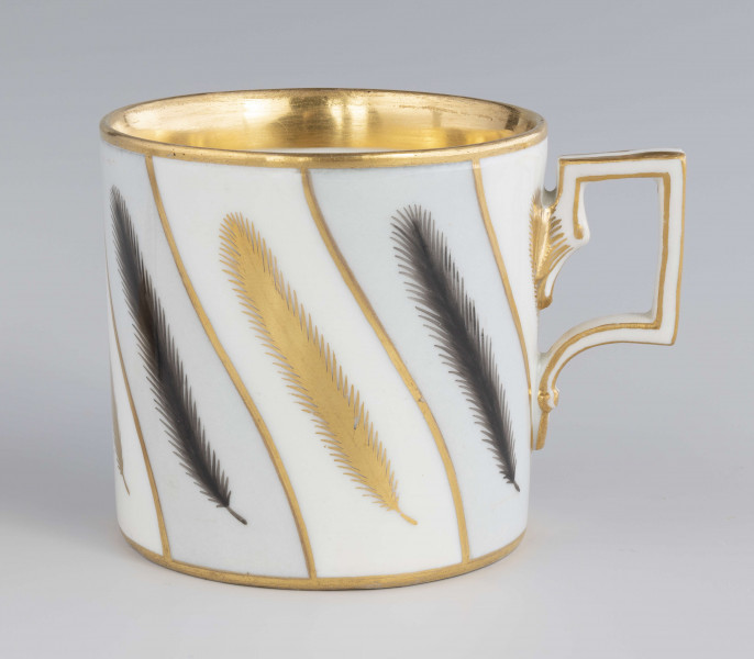 Filiżanka i spodek malowane w piórka. Filiżanka cylindryczna, ucho łamane z listkami w osadzeniach. Brzeg złocony obustronnie, złotem  konturowane ucho, złote kontury dzielą powierzchnię na ukośne pasy na przemian białe i jasnoszare. W nich malowane odpowiednio złote i czarne pióra.