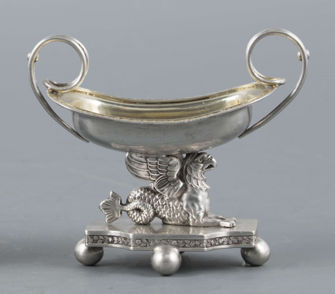 S/Mt/359/ML - Solniczka w kształcie prostokątnej łódki z dwoma wolutowymi uchami z okrągłego drutu, umieszczonej na figurce gryfa, z  podstawą prostokątną o wklęsłych narożach i czterech nóżkach w formie spłaszczonych kulek. Brzeg łódki wywinięty, wnętrze złocone. Brzegi łódki i boki podstawy obiega moletowana taśma z ornamentem roślinnym.