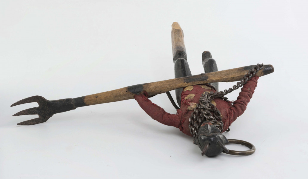 Figurka z szopki, diabeł, rzeźbiona w drewnie. Pomalowana na czarno. Ubrana w czerwony kaftan. Na szyi łańcuch. Do łańcucha doczepione widły, które diabeł trzyma obydwoma rękami. Twarz mocno modelowana, głębokie oczodoły, duże uszy, mała broda wysunięta mocno do przodu. Na głowie małe różki. Z tyłu skórzany ogon.