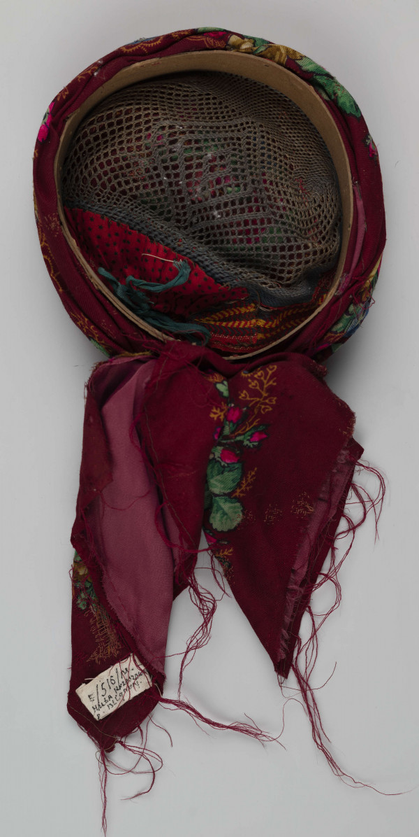 E/516/ML - Czepek mężatki. Denko wewnętrzne wykonane z plecionej siatki zielonej. Z wierzchu chustka fabryczna koloru ciemnoczerwonego w kwiaty wiązana na tekturowej obręczy. Końce chustki zwisają do tyłu.