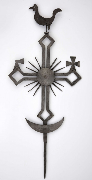 Krzyż żelazny wykonany techniką kucia. Stanowi zakończenie kapliczki przydrożnej. Posiada u góry kogucika, w podstawy - półksiężyce i małe krzyżyki na ramionach. Od skrzyżowania ramion wychodzi 12 promieni.