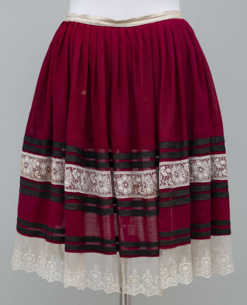 E/897/ML - Zapaska z wełny fabrycznej w kolorze bordowym. U dołu ozdobiona falbaną z białej koronki (szer. 5,5 cm) w odległości 14 cm brzegu i na samym brzegu (szer. 9 cm). Pomiędzy 2 szlaki z 4 czarnych tasiemek oraz nad górną koronką 1 szlak z 2 czarnych tasiemek. Zszyta z 6 równych kawałków tkaniny, na maszynie. Przymarszczona w biały płócienny pasek. 