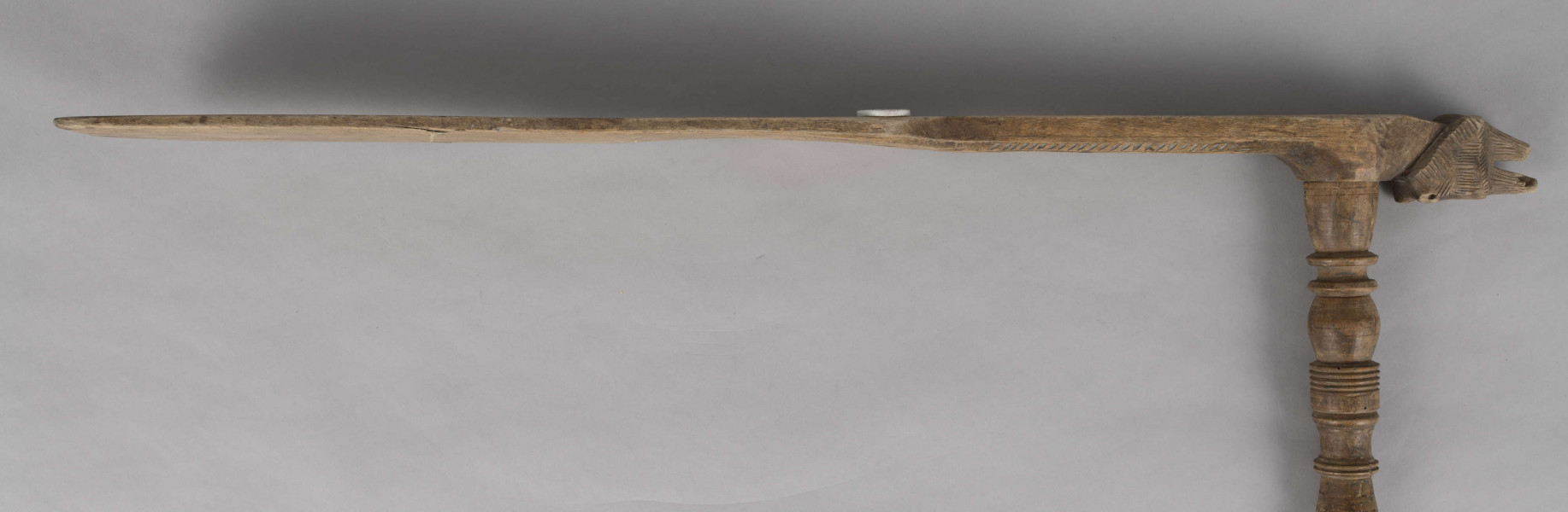 E/8019/ML - Przęślica krężołkowa z drewna brzozowego składa się z cienkiej deski – przysiadki, w której przy jednym końcu osadzona jest pionowa beleczka zw. piórem, z nasadzonym od góry ruchomym krężlem w kształcie wydłużonego stożka. Deseczka przysiadki o kształcie prostokątnym lekko zwężającym, od ok. połowy swej długości w kierunku pióra, i zakończonym głową zwierzęcą – psa. Pióro i krężel ozdobnie toczone. Krężel ma kształt stożka z rozszerzoną podstawą, w której znajduje się stożkowaty otwór, do osadzenia krężla na piórze. Dekoracja snycerska ryta znajduje się na zwężającej części przysiadki. Część zdobiona oddzielona małym stopniem. Grubość przysiadki w części zdobionej 2x większa od pozostałej części. Dekoracja snycerska tworzy ornament roślinny w postaci trzech ulistnionych drobno gałązek. Na zakończeniu ornamentu znajdują się ryte liście oraz inicjały „K.F.”. Głowa psa zdobiona drobnymi nacięcia. Zakończenie przysiadki wycięte faliście.