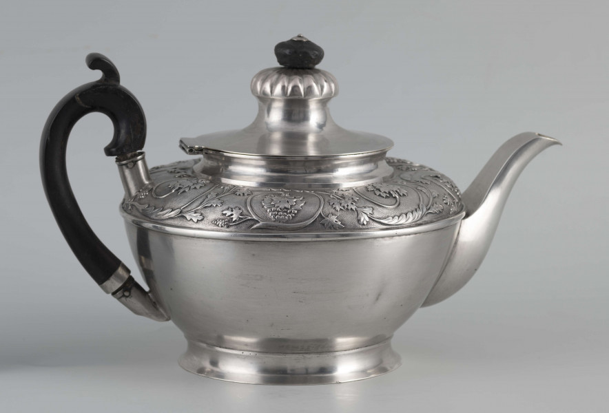 S/Mt/542/ML - Korpus w kształcie spłaszczonej kuli, na niskiej stopie, wylew w formie dzióbka, ucho drewniane malowane na czarno, osadzone w dwóch tulejkach, pokrywka na zawiasie, płaska, w części środkowej wypiętrzona i ozdobiona puklowaną rozetą.Górna część brzuśca ozdobiona sztancowaną taśmą z wzorem spiralnie zwinietej wici roślinnej z kwiatami, uchwyt przykrywki zakończony drewnianą rozetką.  
