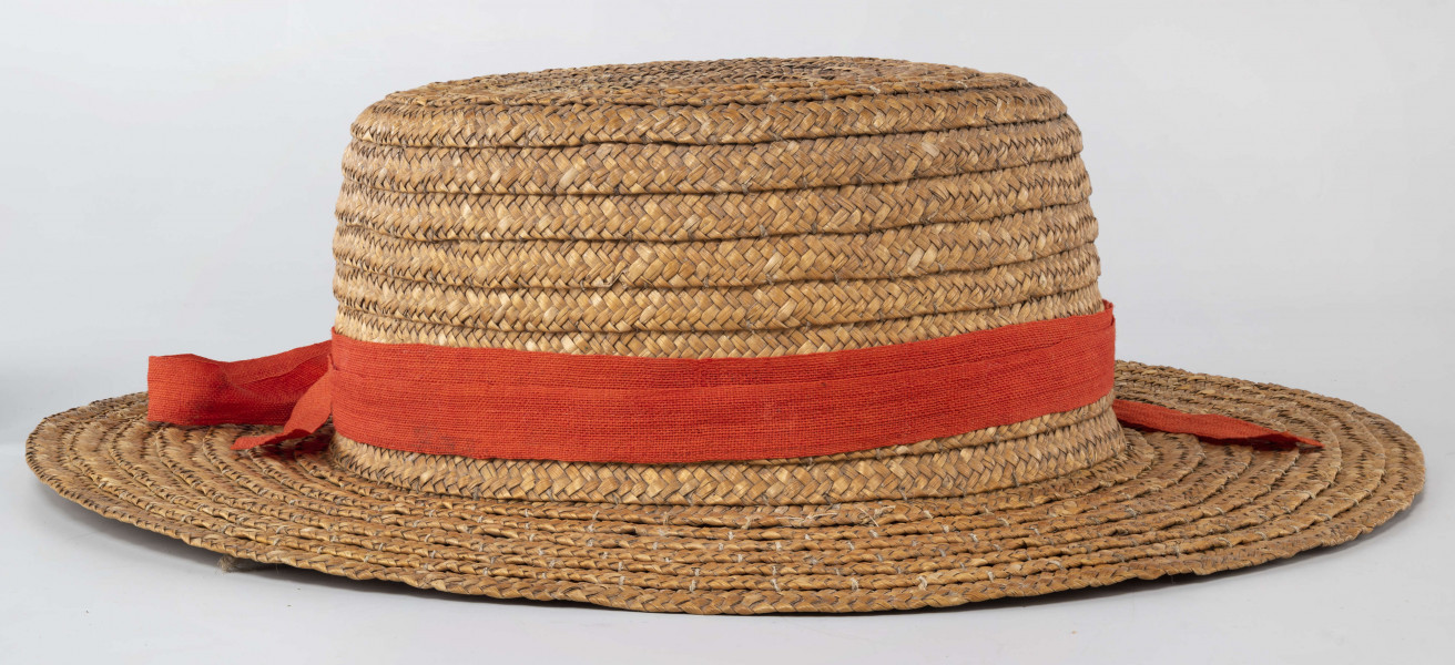 Kapelusz ze słomy owsianej o prostym denku i rondzie płaskim. Upleciony techniką warkoczową. Warkocze bez ząbków, zszyte cienkimi szarymi nićmi. Denko otoczone czerwoną wąską wstążką, zakończone kokardą. 