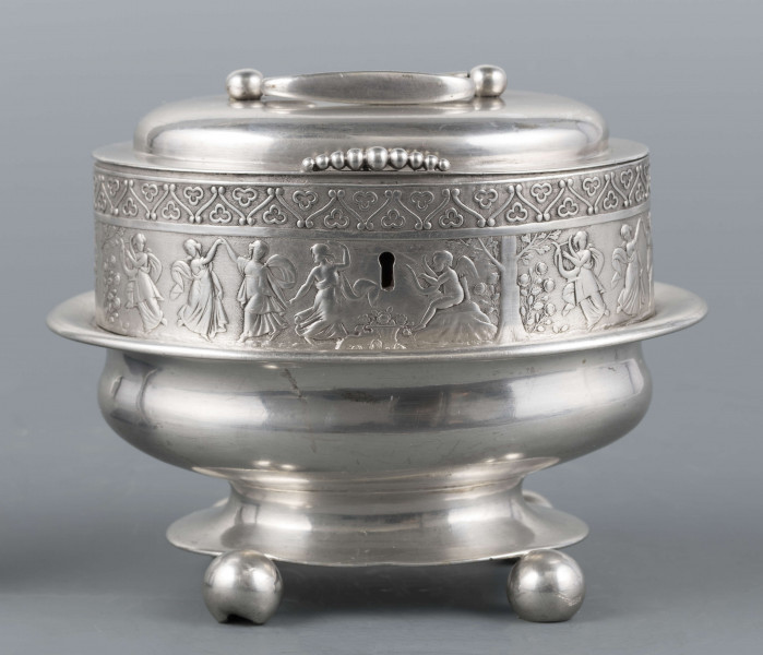 S/Mt/363/ML - Cukiernica owalnana niskiej stopie z czterema kulistymi nóżkami, z pokrywką w formie wieczka.
Podzielona na dwie części : 
- część dolna lekko wybrzuszona, o gładkiej powierzchni, kończy się w połowie wysokości wywiniętym kołnierzem; - część górna prosta uformowana jest z moletowanej taśmy, na której powtarza się trzykrotnie scenka antyczna: po rawej siedzący pod drzewem amorek gra na lirze, po lewej stoi grająca na lirze kobieta, pomiędzy nimi tańczą trzy inne.Górną krzwędź obiega pas stylizowanych neogotyckich trójliści.