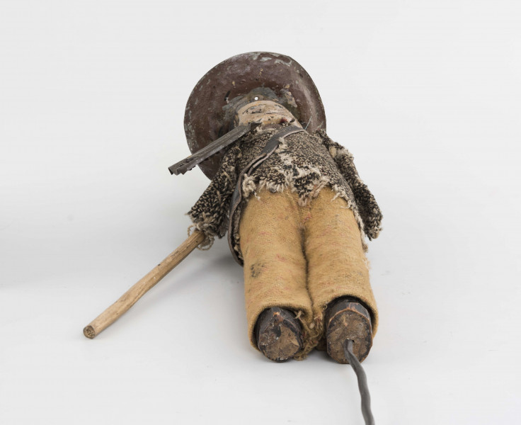 Figurka z szopki - blacharz. Kadłub drewniany na drucie. Twarz lekko ciosana. Rysy twarzy zaznaczony farbą. Blaszany kapelusz, szaro-czarna bluza, rude spodnie. Przez ramię przewieszona torba skórzana na pasku. Na piersi przymocowane narzędzia kuchenne z blachy. W prawym ręku drewniany kij.