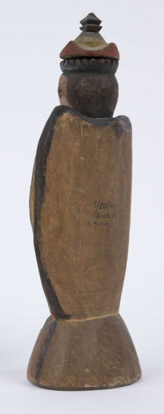 E/8100/ML - Rzeźba pełna wykonana z jednego kawałka drewna lipowego, polichromowana. Postać w hieratycznej postaci, w układzie frontalnym. Ręce wsparte na biodrach. Postać stoi na stożkowatym cokole. Na głowie korona z dwóch oddzielnych kawałków drewna, złączonych ozdobnym czopem drewnianym. Włosy czarne, ręce i twarz różowa, korona żółta z czerwonym obrzeżem, zwieńczenie czarne. Płaszcz stylizowany, przypominający skrzydła lub ornat, czarny. Szaty wewnętrzne żółte z czerwonymi plamkami. Wewnątrz długie, zielone z niebieskimi plamami. Buty czarne. Na piersiach duże otwarte serce zwieńczone czarnym krzyżem. Cokół w naturalnym kolorze drewna, ozdobiony pionowymi niebieskimi paskami i czerwonymi kropkami. Wym. cokołu: 13 x 10,5 cm. Sygnatura na spodzie (pisana): Jan Kaproń.