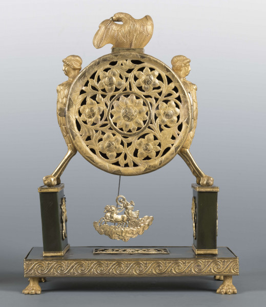 S/Mt/640/ML - Wsparty na czterech zwierzęcych łapach z brązu złoconego, podstawa prostokątna, zdobiona zgeometryzowanym fryzem, wyżej dwa prostokątne filary przechodzące w hybrydyczne figury podtrzymujące cylindryczną puszkę mechanizmu, zwieńczoną figurą orła. Tarcza emaliowana z podziałem arabskim - na niej sygnatura kursywą 