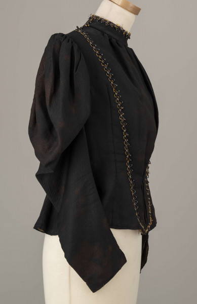 E/8038/ML - Kaftan kobiecy zszyty z 10 kawałków czarnej wełenki. Składa się z 2 części przodu, pleców i rękawów. Wycięcie wokół szyi wykończone stójką szer. 3,5 cm. Przód zapinany z boku na haftki. Rękawy w góry bufiaste, suto marszczone, zwężające się. Przód ułożony w zakładki. Stójka i przód ozdobiony żółtym sznureczkiem, czarną sieczką i białymi szklanymi koralikami. Szyty na maszynie.