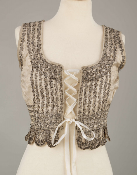 E/6235/ML - Gorset ślubny z białego wzorzystego atłasu. Składa się z 3 części: pleców i dwóch przodów. W pasie doszyte 17 owalnych klapek, zw. taszki. Wycięcie wokół szyi , przodów i rękawów wykończone białą wełnianą tasiemką, zw. szczotka. Całość zdobiona złotym szychem i srebrnymi cekinami. Podszyty białą bawełnianą podszewką.  