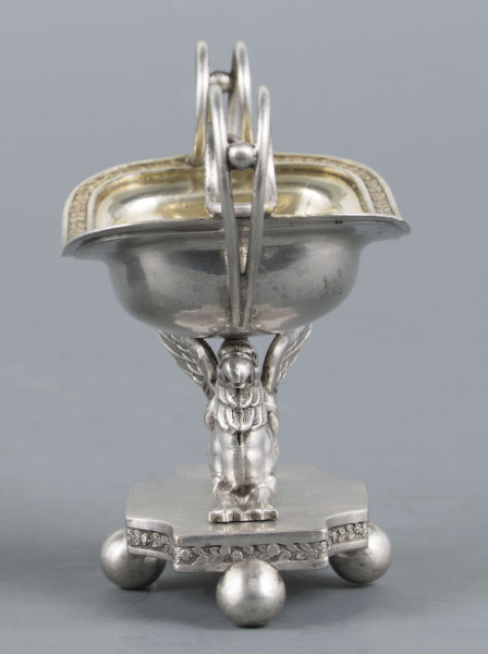 S/Mt/359/ML - Solniczka w kształcie prostokątnej łódki z dwoma wolutowymi uchami z okrągłego drutu, umieszczonej na figurce gryfa, z  podstawą prostokątną o wklęsłych narożach i czterech nóżkach w formie spłaszczonych kulek. Brzeg łódki wywinięty, wnętrze złocone. Brzegi łódki i boki podstawy obiega moletowana taśma z ornamentem roślinnym.