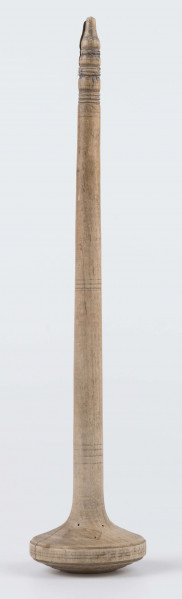 E/8019/ML - Przęślica krężołkowa z drewna brzozowego składa się z cienkiej deski – przysiadki, w której przy jednym końcu osadzona jest pionowa beleczka zw. piórem, z nasadzonym od góry ruchomym krężlem w kształcie wydłużonego stożka. Deseczka przysiadki o kształcie prostokątnym lekko zwężającym, od ok. połowy swej długości w kierunku pióra, i zakończonym głową zwierzęcą – psa. Pióro i krężel ozdobnie toczone. Krężel ma kształt stożka z rozszerzoną podstawą, w której znajduje się stożkowaty otwór, do osadzenia krężla na piórze. Dekoracja snycerska ryta znajduje się na zwężającej części przysiadki. Część zdobiona oddzielona małym stopniem. Grubość przysiadki w części zdobionej 2x większa od pozostałej części. Dekoracja snycerska tworzy ornament roślinny w postaci trzech ulistnionych drobno gałązek. Na zakończeniu ornamentu znajdują się ryte liście oraz inicjały „K.F.”. Głowa psa zdobiona drobnymi nacięcia. Zakończenie przysiadki wycięte faliście.