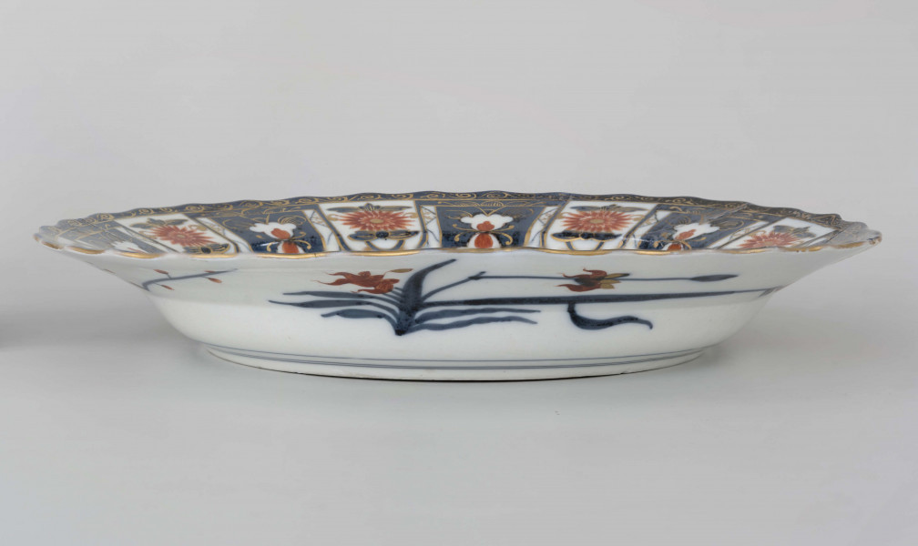 Półmisek okrągły o brzegu falistym. Na kołnierzu szeroka bordiura typu lambrekinowego z dodanymi motywami kwiatowymi, pośrodku dna gałązka kwiatowa, wokół regularnie rozmieszczone mniejsze. Dekoracja dwubarwna: partie kobaltowe malowane pod szkliwem, kwiaty malowane czerwienią naszkliwnie, całość o rysunku podkreślonym złoceniem. Na odwrocie kołnierza malowane gałęzie śliwy i irysów.
 