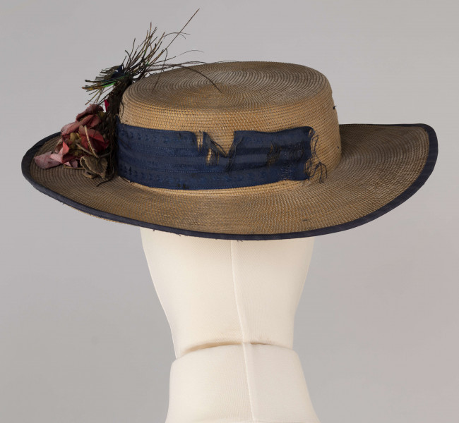 E/560/ML - Kapelusz męski, z cienkiej trzcinki zszywanej nicią. Rondo obszyte niebieską tasiemką, opasane również niebieską (ciemnoniebieską/jasnogranatową) jedwabną wstążką. Kapelusz ozdobiony jest sztucznymi kwiatami białymi i jasnoróżowymi oraz pawimi piórami. Pióra przypiete są metalowo-plastikową broszką.