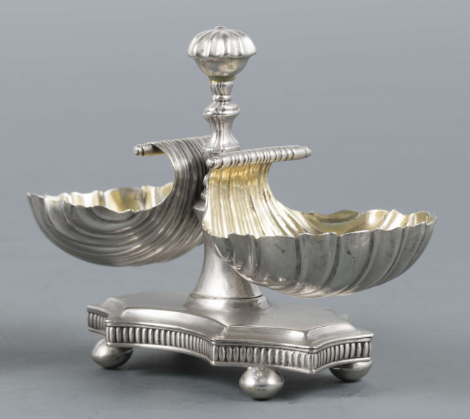 S/Mt/494/ML - Solniczka składa się z dwóch czarek w formie muszli połączonych z pionowym, profilowanym uchwytem o zwieńczniu w formie makówki ustawionym na wydłużonej ośmiobocznej podstawie o wklęsłych bokach. Krawędź podstawy fasetowana, boki karbowane, cztery nóżki w kształcie spłaszczonych kulek. Wnetrze muszli złocone.
Na dnie podstawy sygnatura kursywą w owalnych ramkach:
- J.Gałecki
- w Lublinie
- punca owalna z przedstawieniem dłoni (sygnatura pomocnicza)
- wężyk probierczy
- znak probierni warszawskiej z próbą 