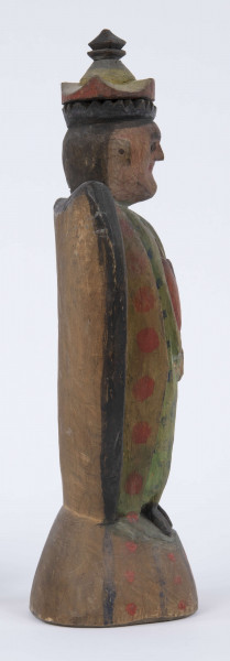 E/8100/ML - Rzeźba pełna wykonana z jednego kawałka drewna lipowego, polichromowana. Postać w hieratycznej postaci, w układzie frontalnym. Ręce wsparte na biodrach. Postać stoi na stożkowatym cokole. Na głowie korona z dwóch oddzielnych kawałków drewna, złączonych ozdobnym czopem drewnianym. Włosy czarne, ręce i twarz różowa, korona żółta z czerwonym obrzeżem, zwieńczenie czarne. Płaszcz stylizowany, przypominający skrzydła lub ornat, czarny. Szaty wewnętrzne żółte z czerwonymi plamkami. Wewnątrz długie, zielone z niebieskimi plamami. Buty czarne. Na piersiach duże otwarte serce zwieńczone czarnym krzyżem. Cokół w naturalnym kolorze drewna, ozdobiony pionowymi niebieskimi paskami i czerwonymi kropkami. Wym. cokołu: 13 x 10,5 cm. Sygnatura na spodzie (pisana): Jan Kaproń.