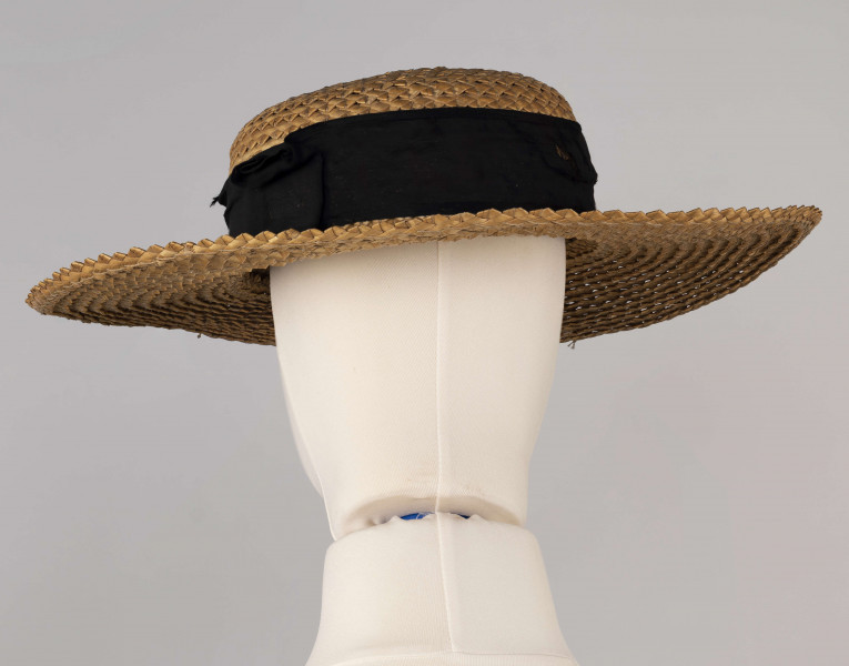 E/586/ML - Kapelusz ze słomy żytniej, o owalnym denku i płaskim rondzie. Upleciony techniką warkoczową - warkocze wąskie, ząbkowane, zszyte szarymi nićmi. Denko otoczone czarną, jedwabną wstążką o szerokości 5 cm.
