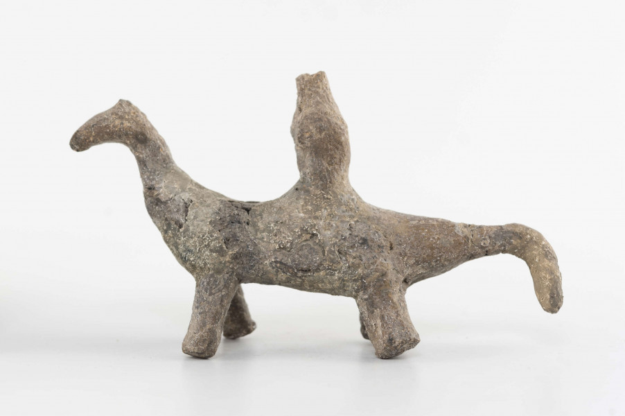 Figurka gliniana jeźdźca na koniu wykonana z wałka glinianego, szyjka wyciagnięta do góry, zwężona, zakończona spłaszczoną głową. Ogon mocno ścieniony, załamany do dołu. Kończyny walcowate, doklejone do korpusu. Postać siedząca na grzbiecie wykonana schematycznie. Surowiec - glina z domieszką piasku. Barwa brunatna. Stan zachowania dobry - drobne ubytki. Zabytek sklejany, uzupełniany gipsem.
Wymiary dodatkowe - 1,9 cm (grub. korpusu); 3 cm (wys. postaci); 1,4 cm (grub. postaci)