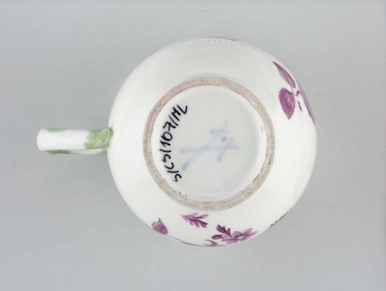 Filiżanka cylindryczna z zaokrąglonym dnem na pierścieniowatej podstawie. Ucho w formie gałązki z listkami przy górnym osadzeniu. Malowana purpurą w kwiaty i owoce (figi ?), ucho podmalowane zielenią.
 