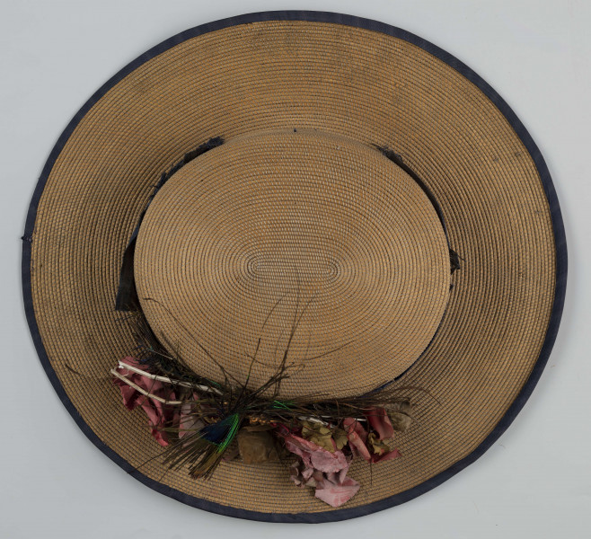 E/560/ML - Kapelusz męski, z cienkiej trzcinki zszywanej nicią. Rondo obszyte niebieską tasiemką, opasane również niebieską (ciemnoniebieską/jasnogranatową) jedwabną wstążką. Kapelusz ozdobiony jest sztucznymi kwiatami białymi i jasnoróżowymi oraz pawimi piórami. Pióra przypiete są metalowo-plastikową broszką.