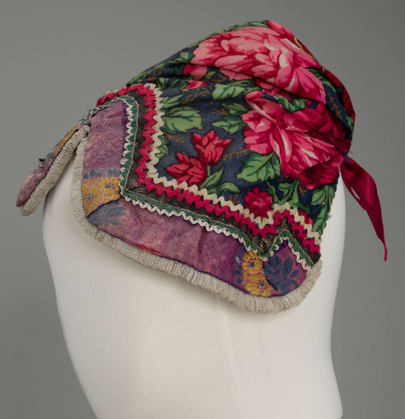 Czepek mężatki na płóciennej podszewce z granatowej cienkiej wełenki w amarantowe róże i jasnozielone listki. Marszczony na środku przodu i tyłu. Przód ozdobionymi kremowymi i amarantowymi ząbkami, srebrną metaliczną wstążką oraz szeroką atłasową wstążką w kolorze fioletowym w granatowe i żółte kwiaty. Brzeg wykończony białą tiulową, drobno plisowaną kryzą. Przód wycięty w duży ząb w kształt płatka kwiatu.