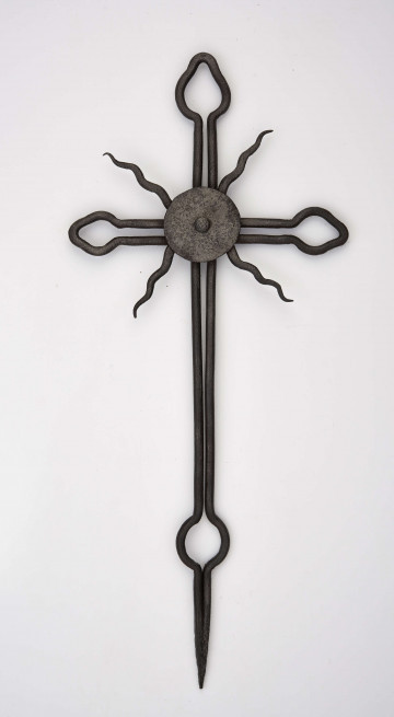 Krzyż wykonany z okrągłego żelaza, technika kucia i wyginania. Całość oprócz promieni wykonana jest z jednego kawałka okrągłego żelaza. W środku, na skrzyżowaniu ramion znajdują się 4 faliste promienie przymocowane dwoma znitowanymi kołami. Służy do ozdoby szczytu drewnianego krzyża.