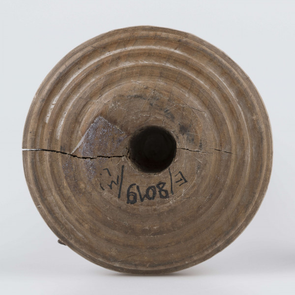 E/8019/ML - Przęślica krężołkowa z drewna brzozowego składa się z cienkiej deski – przysiadki, w której przy jednym końcu osadzona jest pionowa beleczka zw. piórem, z nasadzonym od góry ruchomym krężlem w kształcie wydłużonego stożka. Deseczka przysiadki o kształcie prostokątnym lekko zwężającym, od ok. połowy swej długości w kierunku pióra, i zakończonym głową zwierzęcą – psa. Pióro i krężel ozdobnie toczone. Krężel ma kształt stożka z rozszerzoną podstawą, w której znajduje się stożkowaty otwór, do osadzenia krężla na piórze. Dekoracja snycerska ryta znajduje się na zwężającej części przysiadki. Część zdobiona oddzielona małym stopniem. Grubość przysiadki w części zdobionej 2x większa od pozostałej części. Dekoracja snycerska tworzy ornament roślinny w postaci trzech ulistnionych drobno gałązek. Na zakończeniu ornamentu znajdują się ryte liście oraz inicjały „K.F.”. Głowa psa zdobiona drobnymi nacięcia. Zakończenie przysiadki wycięte faliście.