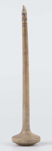 E/8019/ML - Przęślica krężołkowa z drewna brzozowego składa się z cienkiej deski – przysiadki, w której przy jednym końcu osadzona jest pionowa beleczka zw. piórem, z nasadzonym od góry ruchomym krężlem w kształcie wydłużonego stożka. Deseczka przysiadki o kształcie prostokątnym lekko zwężającym, od ok. połowy swej długości w kierunku pióra, i zakończonym głową zwierzęcą – psa. Pióro i krężel ozdobnie toczone. Krężel ma kształt stożka z rozszerzoną podstawą, w której znajduje się stożkowaty otwór, do osadzenia krężla na piórze. Dekoracja snycerska ryta znajduje się na zwężającej części przysiadki. Część zdobiona oddzielona małym stopniem. Grubość przysiadki w części zdobionej 2x większa od pozostałej części. Dekoracja snycerska tworzy ornament roślinny w postaci trzech ulistnionych drobno gałązek. Na zakończeniu ornamentu znajdują się ryte liście oraz inicjały „K.F.”. Głowa psa zdobiona drobnymi nacięcia. Zakończenie przysiadki wycięte faliście.