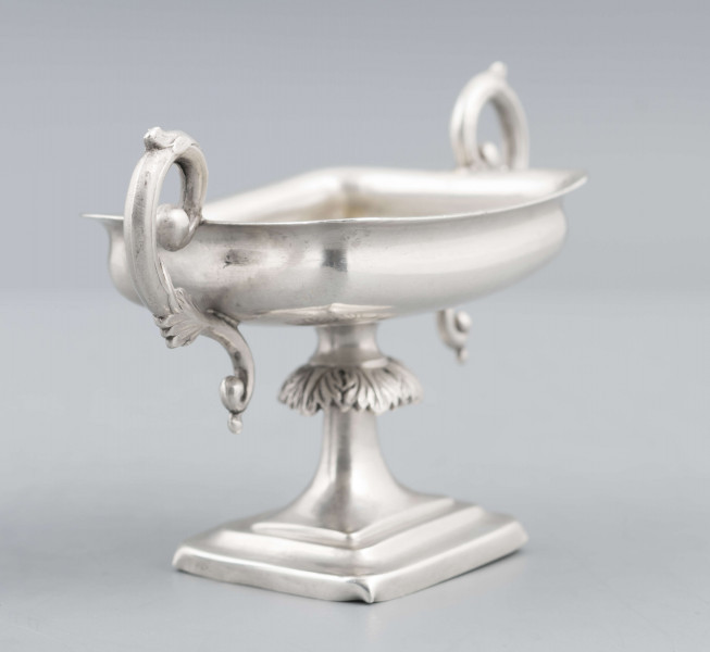 S/Mt/57/ML - Stopa prostokątna, wysklepiona, przechodząca w trzon z owalnym kapturkiem sztancowanym we wzór akantu. Czarka prostokatna o zaokrąglonych narożach i wywinietym brzegu, z dwoma uchami na krótszych bokach. Ucha wolutowe zdobione listkami. 