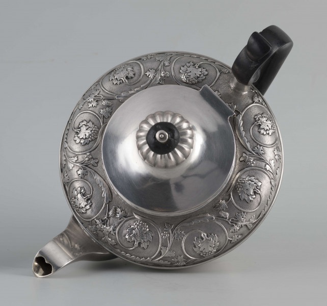 S/Mt/542/ML - Korpus w kształcie spłaszczonej kuli, na niskiej stopie, wylew w formie dzióbka, ucho drewniane malowane na czarno, osadzone w dwóch tulejkach, pokrywka na zawiasie, płaska, w części środkowej wypiętrzona i ozdobiona puklowaną rozetą.Górna część brzuśca ozdobiona sztancowaną taśmą z wzorem spiralnie zwinietej wici roślinnej z kwiatami, uchwyt przykrywki zakończony drewnianą rozetką.  
