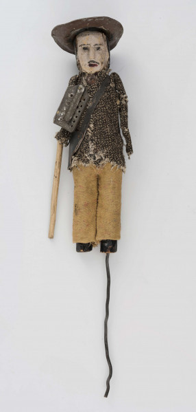 Figurka z szopki - blacharz. Kadłub drewniany na drucie. Twarz lekko ciosana. Rysy twarzy zaznaczony farbą. Blaszany kapelusz, szaro-czarna bluza, rude spodnie. Przez ramię przewieszona torba skórzana na pasku. Na piersi przymocowane narzędzia kuchenne z blachy. W prawym ręku drewniany kij.