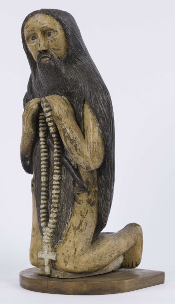 E/6353/ML - Figura drewniana, zakomponowana na postumencie, polichromowana. Postać klęcząca przedstawia św. Onufrego. Twarz płaska, trójkątna, głęboko osadzone oczy, czarne źrenice. Szerokie łuki brwiowe łączą się z wydatnym prostym nosem. Wystające kości jarzmowe. Małe usta otacza bujny zarost: broda i wąsy łączące się z bujnymi włosami, otaczającymi całą postać – na podobieństwo płaszcza. Ręce zgięte w łokciach, uniesione ku górze, łączą się końcami palców w geście modlitwy. W nieproporcjonalnie dużych dłoniach biały różaniec zakończony dużym krzyżem. Nogi masywne z dużymi płaskimi stopami. Prawą dłonią podtrzymuje sękaty kostur. W partii pasa przepasany powrozem z węzłami. 