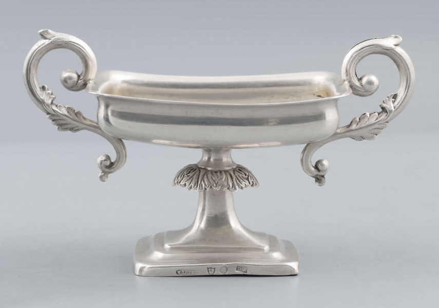 S/Mt/57/ML - Stopa prostokątna, wysklepiona, przechodząca w trzon z owalnym kapturkiem sztancowanym we wzór akantu. Czarka prostokatna o zaokrąglonych narożach i wywinietym brzegu, z dwoma uchami na krótszych bokach. Ucha wolutowe zdobione listkami. 